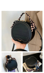 Black round handbag with checkered handle held by a hand, displayed against a neutral background.
